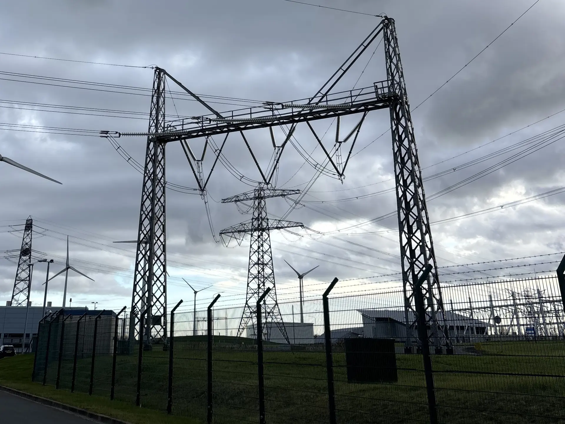 High-voltage pylons and a substation fence with wind turbines behind